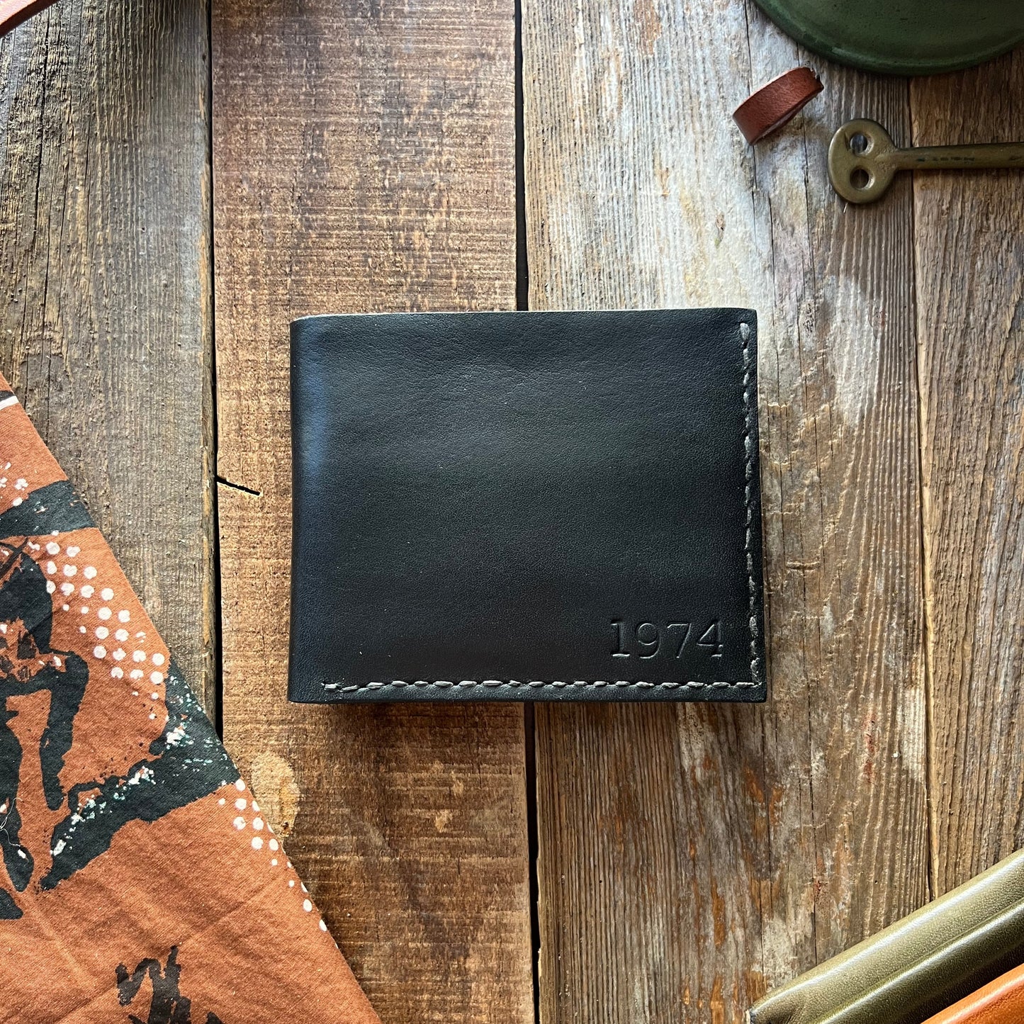Black leather wallet with '1974' engraving on a wooden surface with other leather items.