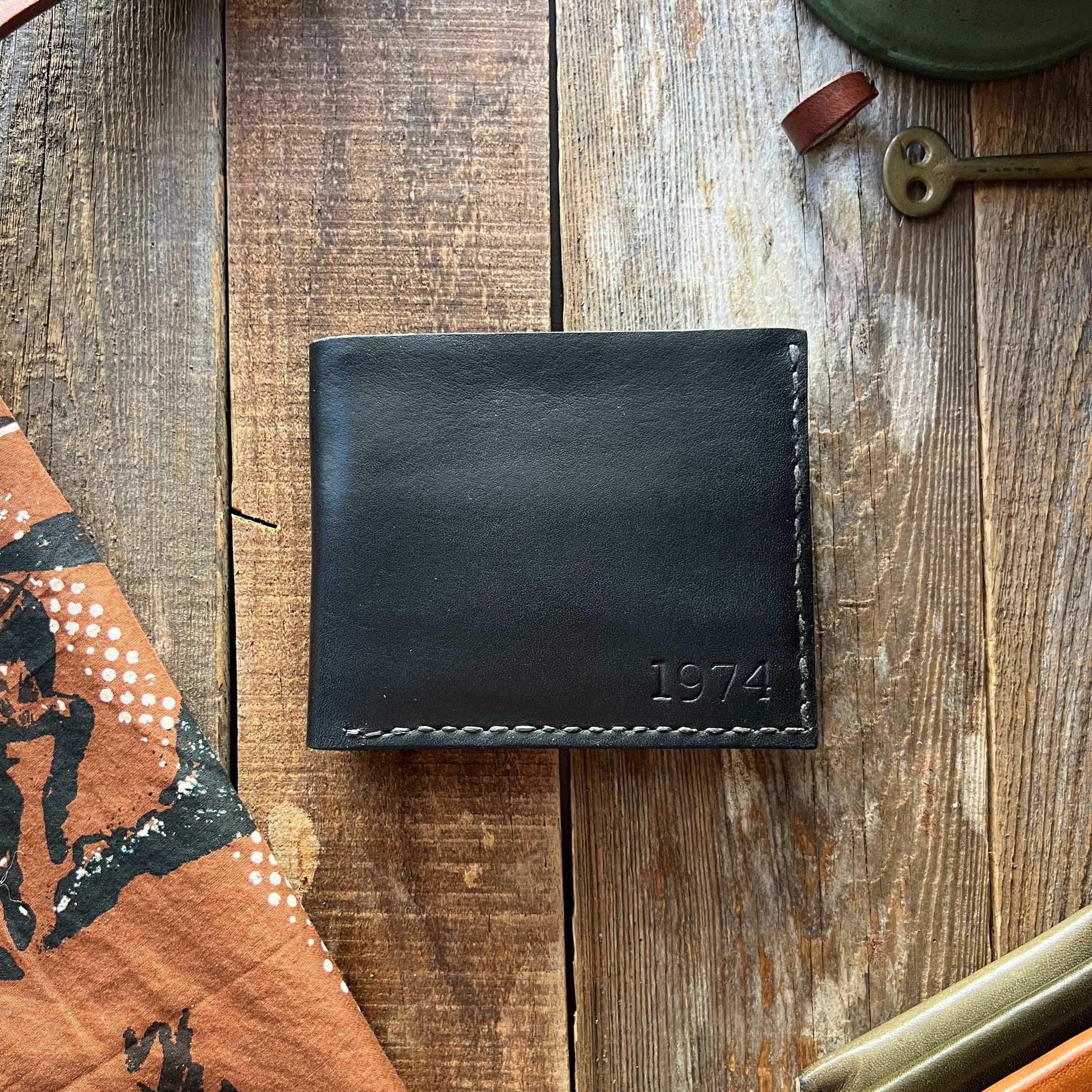 Black leather wallet with '1974' engraving on a wooden surface with other leather items.