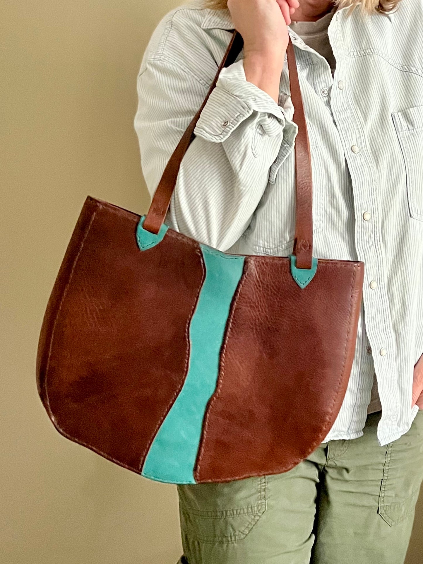 Brown leather tote bag with turquoise stripe held by a person against a beige background