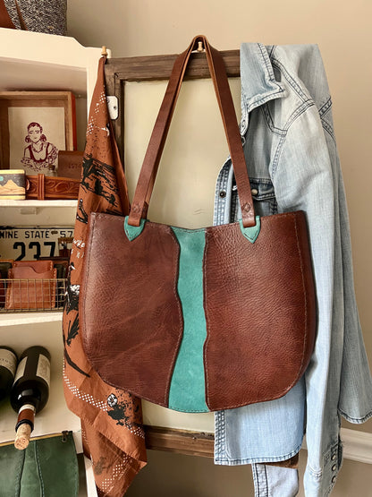 Brown leather tote bag with turquoise accents hanging on a wooden frame against a neutral wall.