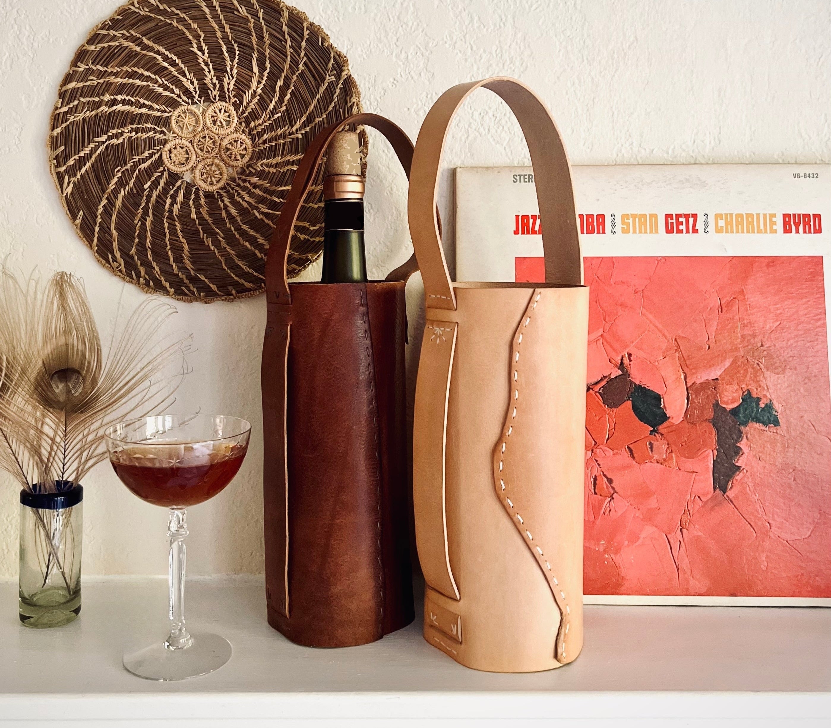Two leather wine bottle bags, one dark brown and one light tan, displayed on a white shelf next to a wine glass, a woven wall hanging, and a vinyl record.