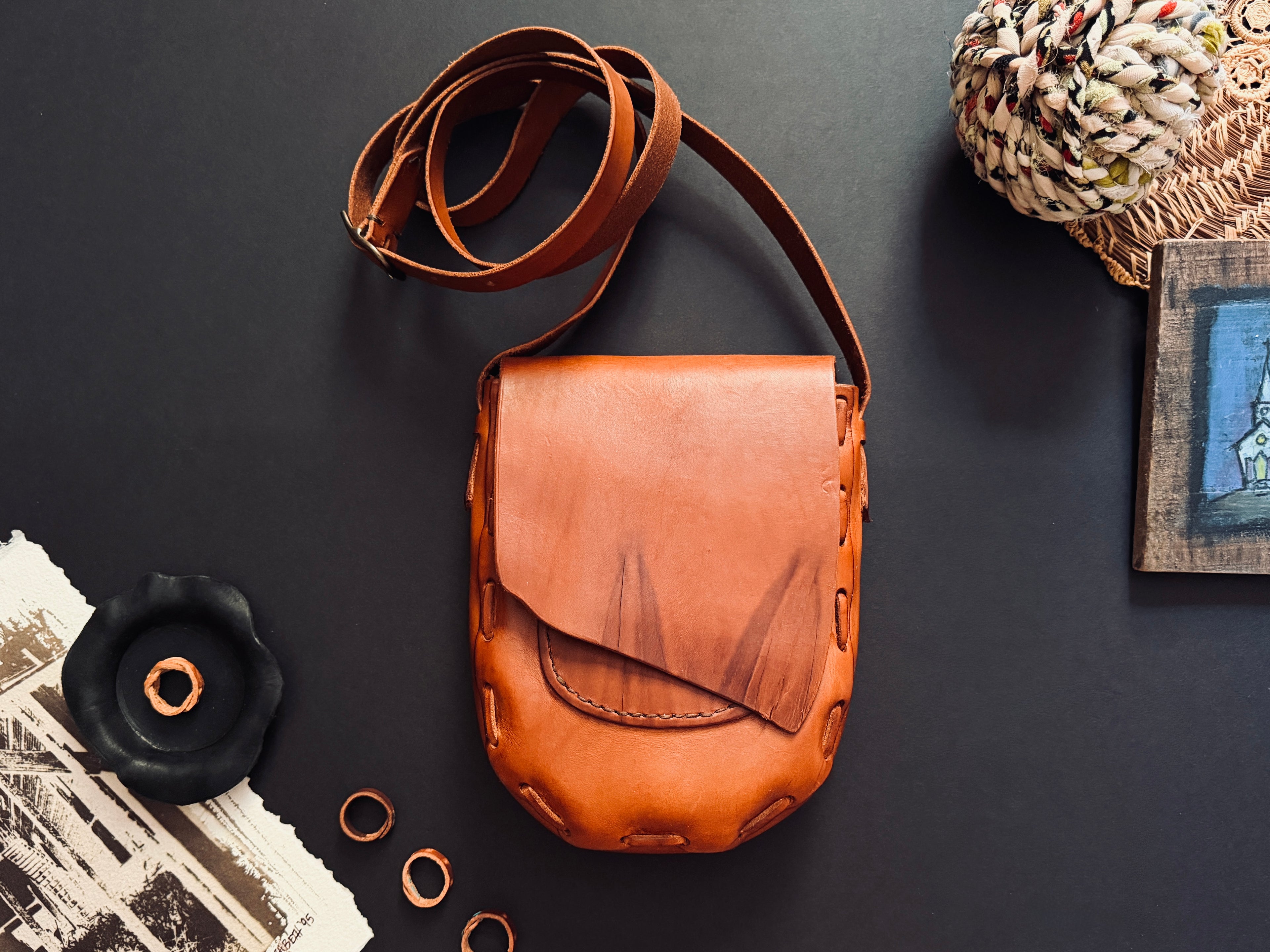 A brown leather 1974 Canyon Bag crossbody bag with a long strap, laid flat on a dark surface with decorative props including rings, a black flower-shaped object, a woven textile, and a small framed print.