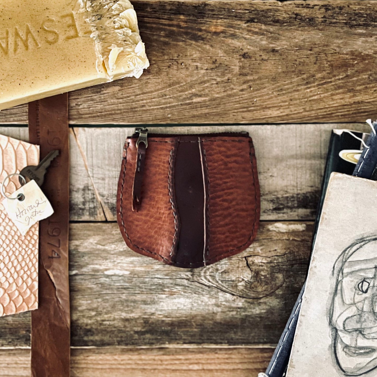 image of 1974s Little River Coin Pouch in Hickory and Chestnut leather resting on a rustic wooden backdrop with beeswax, leather strips and illustrations are surrounding the pouch.