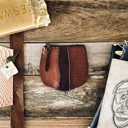 image of 1974s Little River Coin Pouch in Hickory and Chestnut leather resting on a rustic wooden backdrop with beeswax, leather strips and illustrations are surrounding the pouch.