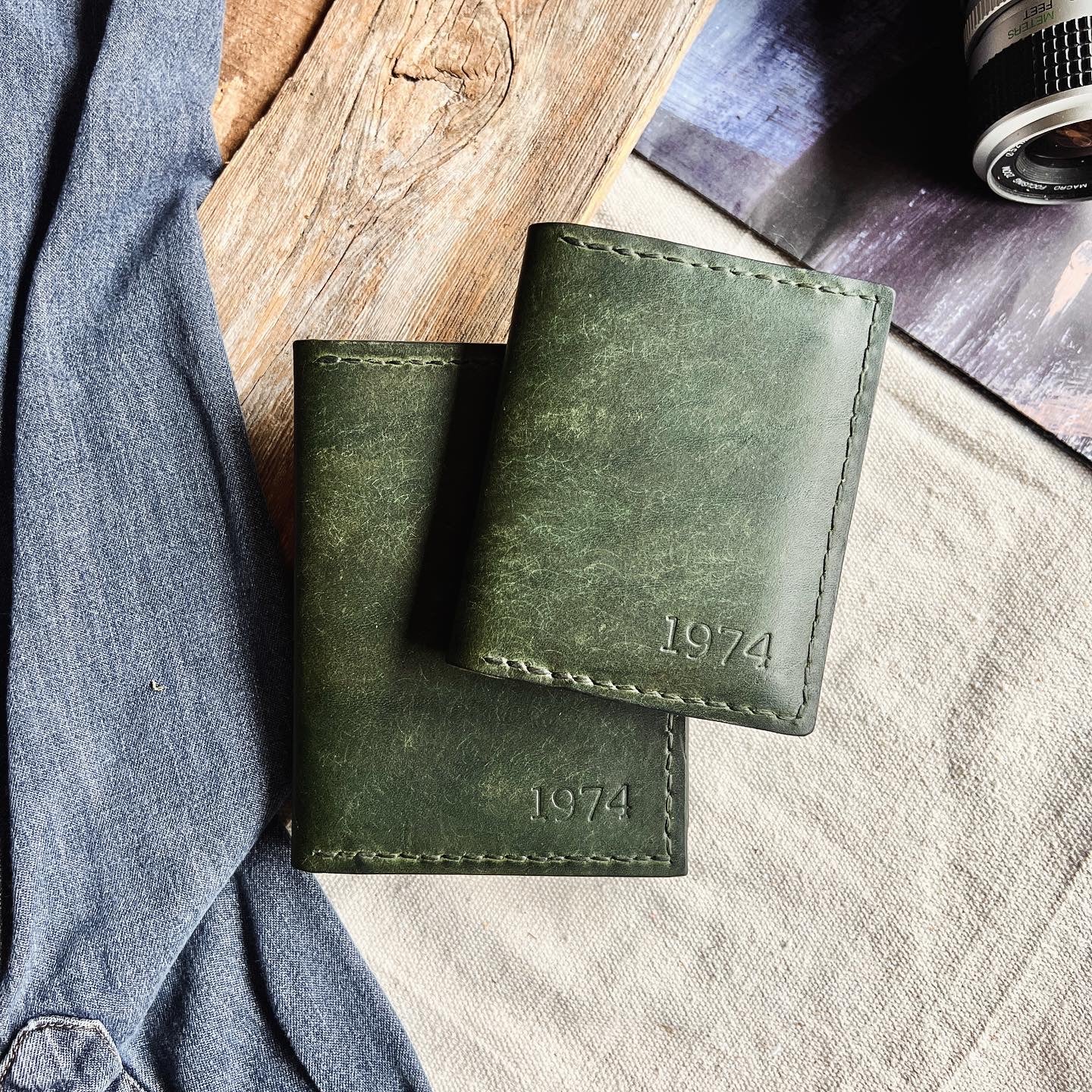 Two artisan made leather wallets on a layered and textured background of woods and canvas with denim and an old camera within view.