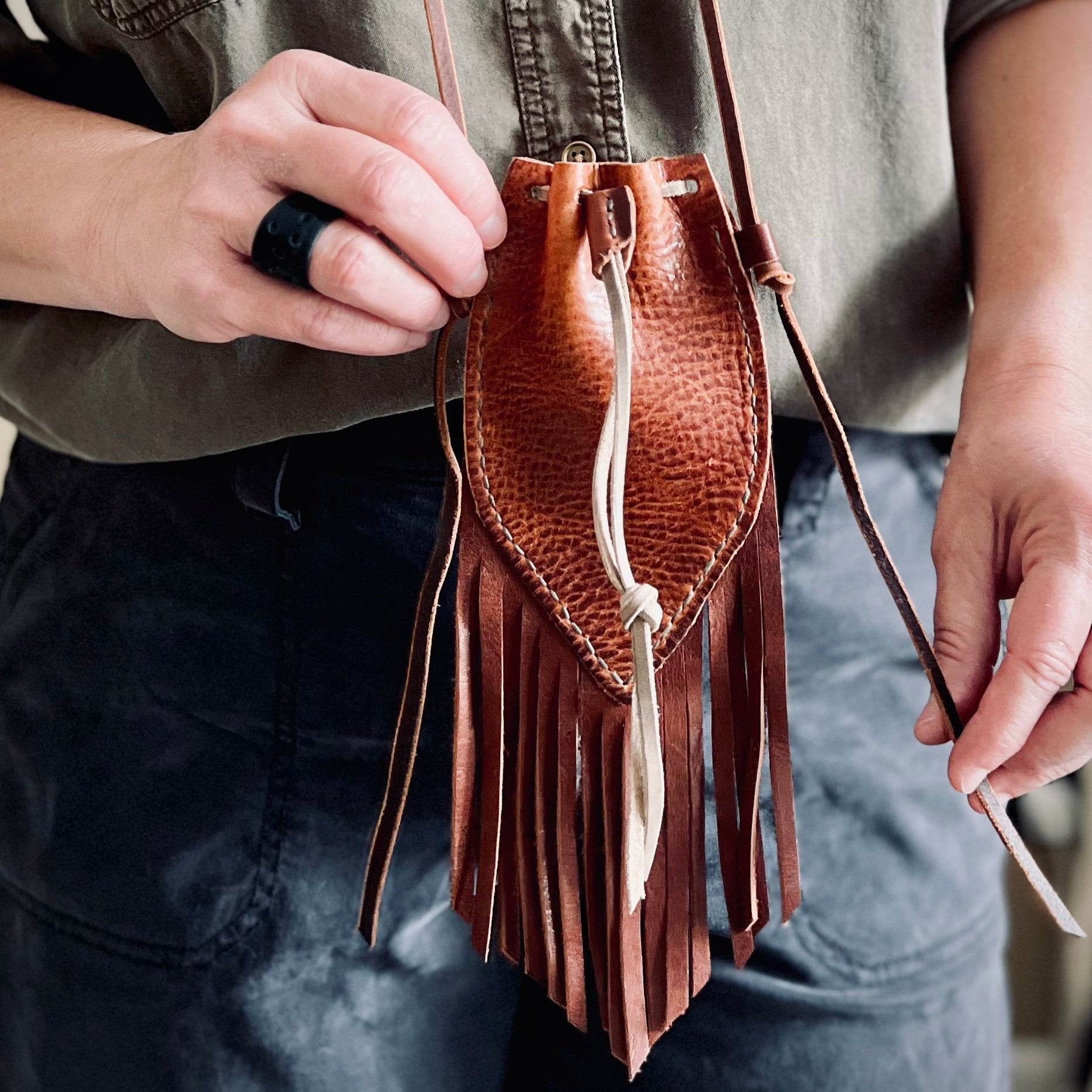 Brown leather fringe bag held by a person wearing a green shirt and blue jeans.