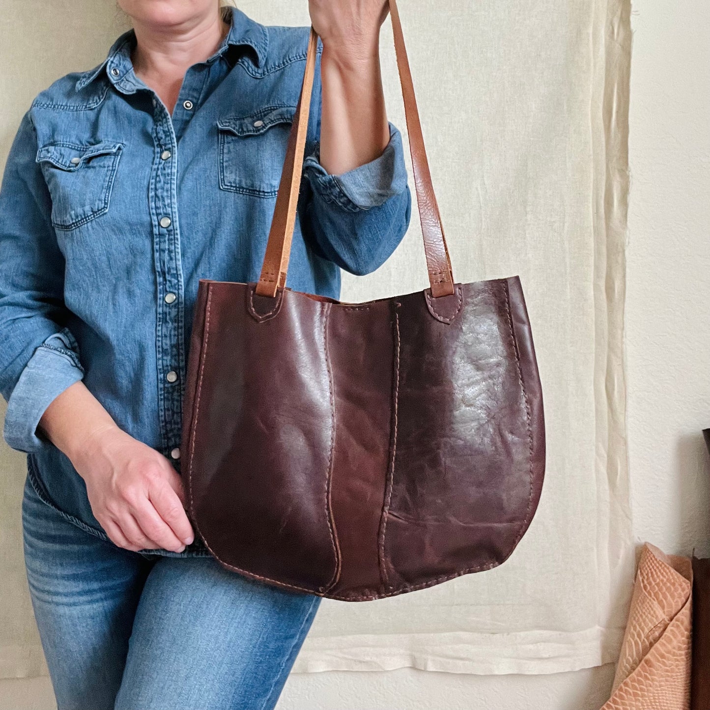 A beautifully rich handmade leather tote bag in chestnut brown held by a woman wearing all denim standing against a beige background with leather hides rolled in the bottom right corner.