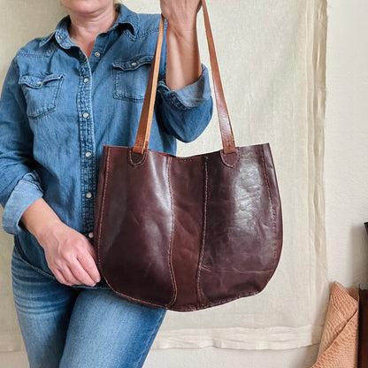 A beautifully rich handmade leather tote bag in chestnut brown held by a woman wearing all denim standing against a beige background with leather hides rolled in the bottom right corner.